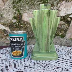 May include: A green ceramic vase with a geometric design. The vase is sitting on a white crocheted doily. A can of Heinz baked beans is in the foreground.