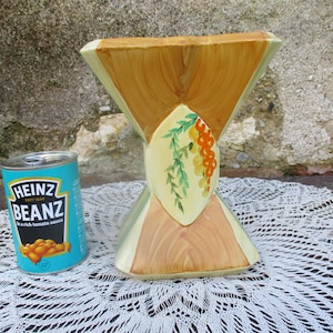 Può includere: Un vaso in ceramica con una forma a clessidra unica e un design dipinto di foglie verdi e bacche arancioni. Il vaso è decorato con un motivo a venature del legno e ha una gamma di colori giallo chiaro, verde e marrone.