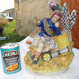 Könnte beinhalten: Eine Keramikfigur eines Mannes in einem Kilt und einem Federhut, der ein Gewehr und eine Trommel hält. Die Figur sitzt auf einem Sockel mit einem gelben und braunen Design. Die Figur ist in leuchtenden Farben bemalt, darunter Rot, Blau, Grün und Gelb.