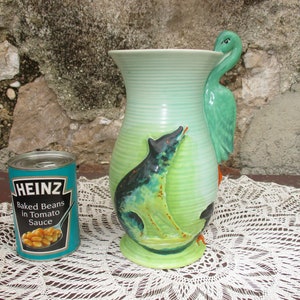 Puede incluir: Un jarrón de cerámica verde y azul con un asa de pájaro y un diseño pintado de un lobo y un pájaro. El jarrón está sobre un mantel de encaje blanco.