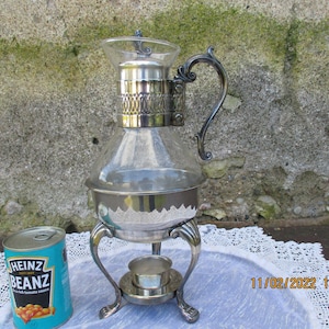 Puede incluir: Tetera de vidrio plateada con tapa de vidrio y soporte de quemador. La tetera tiene un asa decorativa y una banda plateada alrededor del medio. El soporte de quemador tiene tres patas y una taza pequeña en el centro.