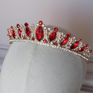 Puede incluir: Una tiara dorada con piedras rojas y transparentes. La tiara tiene forma de corona puntiaguda y está decorada con una fila de piedras rojas.