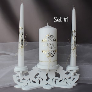 Puede incluir: Conjunto de velas de unidad de boda blancas con un soporte decorativo. La vela de unidad tiene un diseño dorado de "Mr & Mrs". Las dos velas cónicas tienen diseños dorados y están atadas con cuerda.