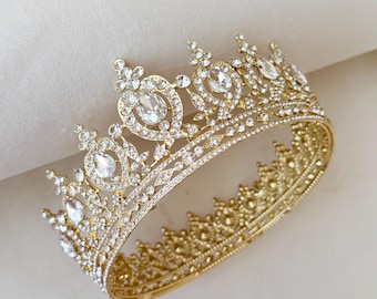 May include: A gold-coloured crown adorned with clear, sparkling rhinestones. The crown features large, oval-shaped rhinestones and smaller, round ones. The base of the crown has a detailed, ornate design.