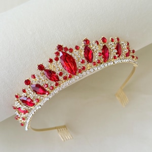 Könnte beinhalten: Ein goldfarbenes Diadem mit einer Reihe von marquise-geschliffenen roten Kristallen in kronenartiger Anordnung. Die Basis ist mit kleinen klaren Strasssteinen besetzt und verfügt über zwei kleine Metallkämme an den Enden für den Halt im Haar.
