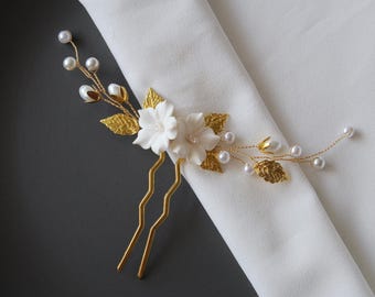 Puede incluir: Dos horquillas doradas con flores blancas y detalles de perlas. Las horquillas están decoradas con hojas doradas y perlas.