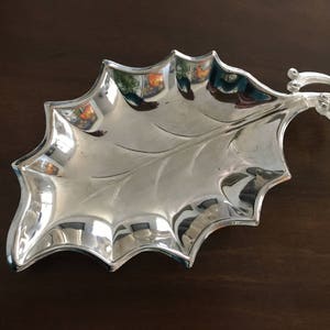 May include: A silver serving dish shaped like a holly leaf with a decorative handle. The dish is highly polished and reflects light.