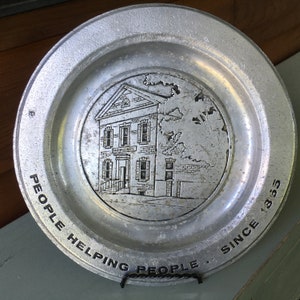 May include: A silver metal plate with a raised image of a building with the text "Mutual Bank" above the entrance. The plate has the text "People Helping People. Since 1855" around the edge.