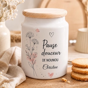 Peut inclure: Un pot en céramique blanc avec un couvercle en bois clair, orné d'un dessin au trait noir représentant des fleurs sauvages, un petit cœur et le texte "Pause douceur DE NOUNOU Christine" imprimé en noir avec des touches rose pâle.