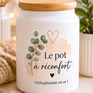 Peut inclure: Un pot de rangement en céramique blanche avec un couvercle en bois, orné d'une branche d'eucalyptus verte, d'un cœur doré et d'un petit cœur noir. Le texte indique Le pot à réconfort et Orthophoniste en or en écriture noire.