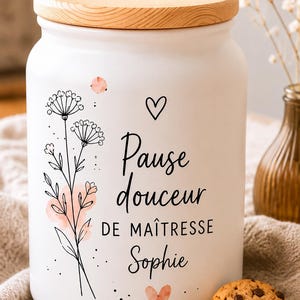 Könnte beinhalten: Ein weißes Keramik-Vorratsglas mit einem hellen Holzdeckel, verziert mit einer schwarzen Strichzeichnung von Wildblumen, einem kleinen Herz und dem Text "Pause douceur DE MAÎTRESSE Sophie" in schwarzer Schrift, akzentuiert durch zarte rosa Aquarellflecken.