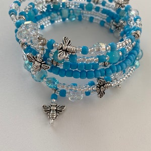 Peut inclure: Un bracelet multi-rangs composé de perles bleues et argentées avec des breloques d'abeilles argentées. Le bracelet est enroulé sur lui-même plusieurs fois.
