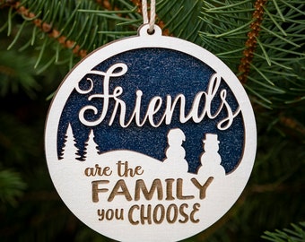 May include: A white wooden Christmas ornament with a blue background. The ornament features the text "Friends are the family you choose" and a silhouette of two snowmen.