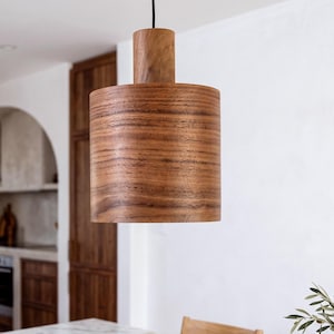Peut inclure: Une suspension cylindrique fabriquée en bois naturel avec un grain visible. La lampe est suspendue par un cordon noir unique et présente une section supérieure cylindrique plus petite qui se connecte au corps principal de l'abat-jour.