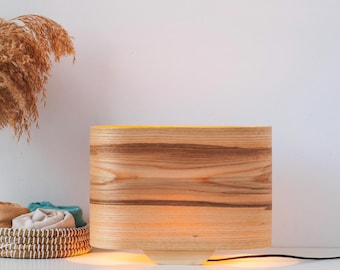 May include: A wooden table lamp with a natural wood grain finish. The lamp has a rounded, oval-shaped shade and a light brown base.