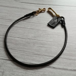 May include: A black leather cord lanyard featuring a gold-toned metal lobster clasp and a hook. A small rectangular black leather tag is attached to the hook, embossed with a graphic design of a pine tree. The cord is arranged in a circular loop.