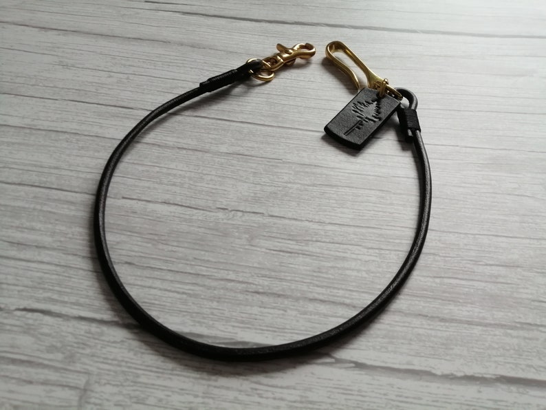 May include: A black leather cord lanyard featuring a gold-toned metal lobster clasp and a hook. A small rectangular black leather tag is attached to the hook, embossed with a graphic design of a pine tree. The cord is arranged in a circular loop.