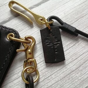 May include: A dark brown leather accessory featuring a gold-toned metal hook and a swivel lobster clasp. A small rectangular dark brown leather tag is attached to the hook, embossed with a stylized tree graphic. A black cord is also connected.