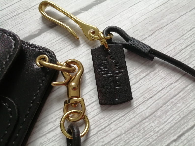 May include: A dark brown leather accessory featuring a gold-toned metal hook and a swivel lobster clasp. A small rectangular dark brown leather tag is attached to the hook, embossed with a stylized tree graphic. A black cord is also connected.