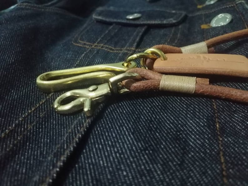 Puede incluir: Primer plano de un gancho y cierre de llave de lat&oacute;n con correas de cuero marr&oacute;n y una peque&ntilde;a etiqueta de cuero. El llavero est&aacute; sobre un fondo de mezclilla azul oscuro.