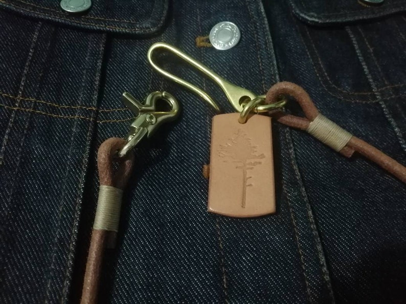 Puede incluir: Primer plano de un llavero de cuero con un cierre de lat&oacute;n y una etiqueta rectangular con un dise&ntilde;o de &aacute;rbol. El llavero es marr&oacute;n con un gancho de lat&oacute;n, sobre una chaqueta vaquera.