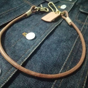 Puede incluir: Una cadena de billetera de cuero marr&oacute;n con un cierre de lat&oacute;n y una peque&ntilde;a etiqueta de cuero. La cadena est&aacute; en bucle y descansa sobre una chaqueta vaquera azul, mostrando el dise&ntilde;o y la textura de la cadena.