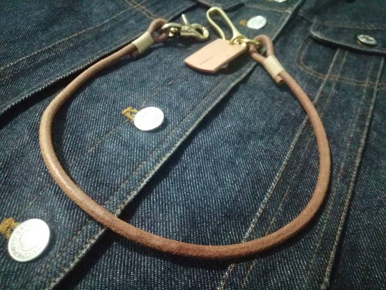 Puede incluir: Una cadena de billetera de cuero marr&oacute;n con un cierre de lat&oacute;n y una peque&ntilde;a etiqueta de cuero. La cadena est&aacute; en bucle y descansa sobre una chaqueta vaquera azul, mostrando el dise&ntilde;o y la textura de la cadena.