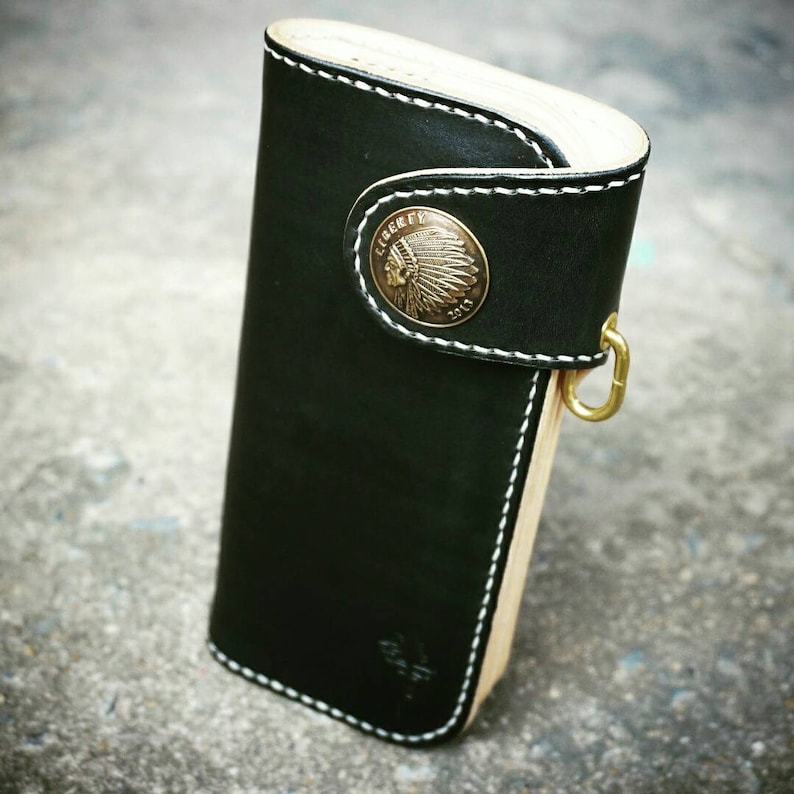 K&ouml;nnte beinhalten: Eine schwarze Ledergeldb&ouml;rse mit wei&szlig;en N&auml;hten und einem Messing-Indianerkopf-Emblem. Die Geldb&ouml;rse hat eine Holzr&uuml;ckseite und einen Messinghaken.