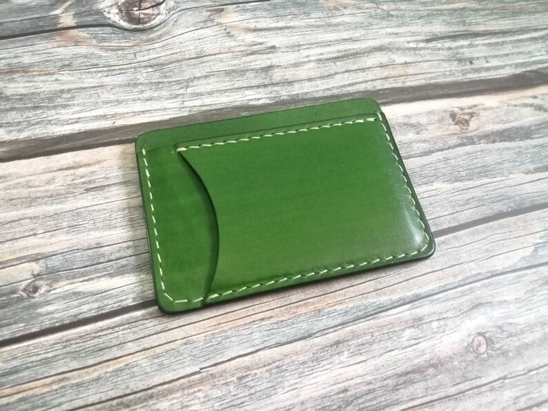 Puede incluir: Un porta tarjetas de cuero verde con costuras blancas. El porta tarjetas tiene un solo bolsillo para tarjetas.