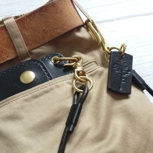 May include: A black leather wallet with a brass snap button, attached to a brass swivel hook and a long black leather cord. A rectangular black leather tag with a stamped tree design hangs from the brass hardware, all secured to tan trousers.