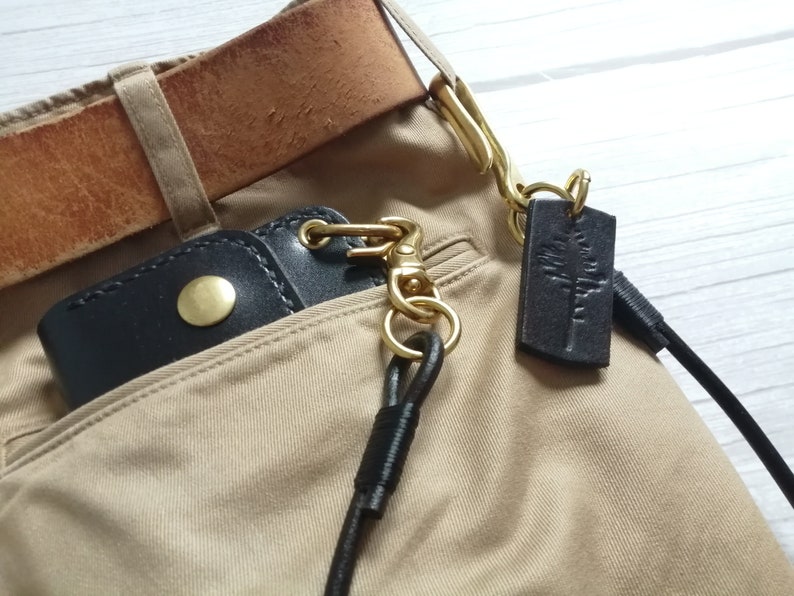 May include: A black leather wallet with a brass snap button, attached to a brass swivel hook and a long black leather cord. A rectangular black leather tag with a stamped tree design hangs from the brass hardware, all secured to tan trousers.
