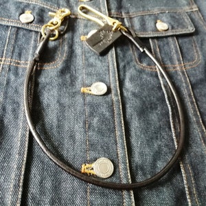 May include: A dark brown leather wallet chain featuring brass hardware, including a swivel lobster clasp and a hook. A small, rectangular black leather tag with an embossed tree design is attached to the brass hook, resting on a dark blue denim jacket.
