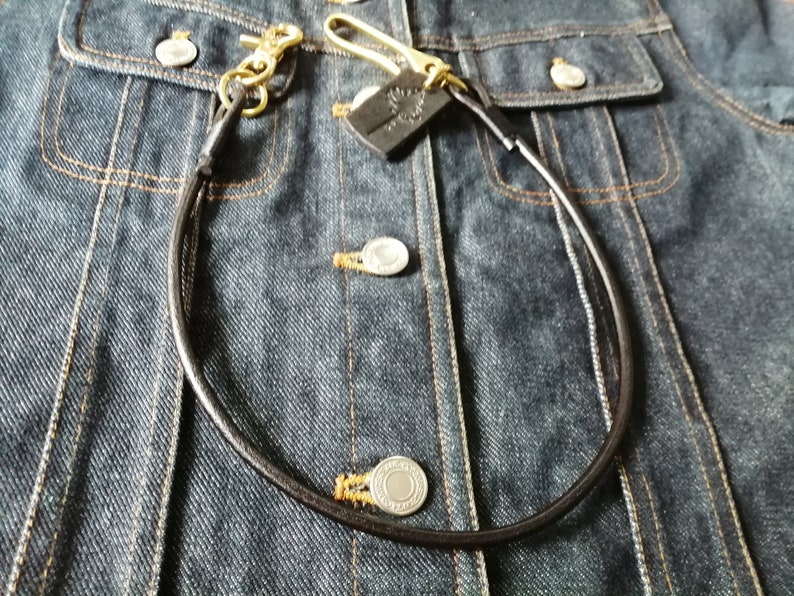 May include: A dark brown leather wallet chain featuring brass hardware, including a swivel lobster clasp and a hook. A small, rectangular black leather tag with an embossed tree design is attached to the brass hook, resting on a dark blue denim jacket.