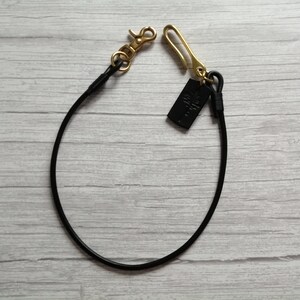 May include: A black leather cord lanyard featuring a gold-toned metal lobster clasp on one end and a gold-toned metal hook on the other. A small, rectangular black leather tag with an embossed tree design is attached to the gold hook.