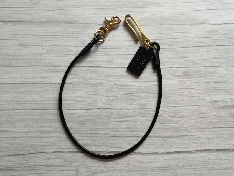 May include: A black leather cord lanyard featuring a gold-toned metal lobster clasp on one end and a gold-toned metal hook on the other. A small, rectangular black leather tag with an embossed tree design is attached to the gold hook.