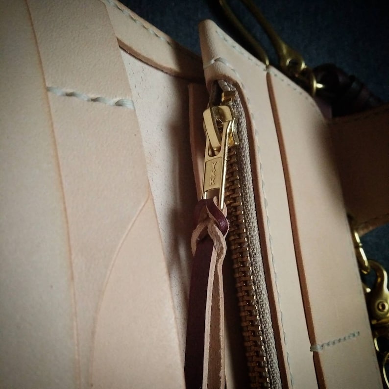 K&ouml;nnte beinhalten: Nahaufnahme eines Lederportemonnaies mit einem goldenen Rei&szlig;verschluss und einem braunen Leder-Zugband. Der Rei&szlig;verschluss tr&auml;gt den Schriftzug "YKK".