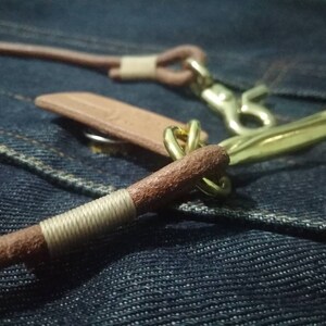 Puede incluir: Primer plano de un cord&oacute;n de cuero con herrajes de lat&oacute;n. El cord&oacute;n de cuero marr&oacute;n tiene una secci&oacute;n envuelta y un cierre de lat&oacute;n. El fondo es una tela vaquera azul texturizada.