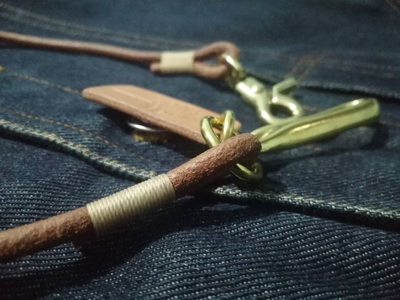 Puede incluir: Primer plano de un cord&oacute;n de cuero con herrajes de lat&oacute;n. El cord&oacute;n de cuero marr&oacute;n tiene una secci&oacute;n envuelta y un cierre de lat&oacute;n. El fondo es una tela vaquera azul texturizada.