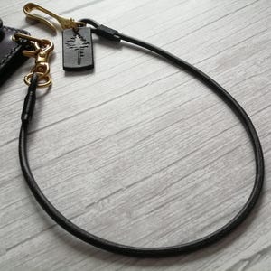 Könnte beinhalten: Eine schwarze Geldbörsenkette aus Leder mit einem Messinghaken und einem kleinen rechteckigen Lederanhänger, der mit einer Tannenbaumgrafik geprägt ist. Die Kette ist an einer schwarzen Lederecke mit Messingdruckknopf und Messingkarabiner befestigt.