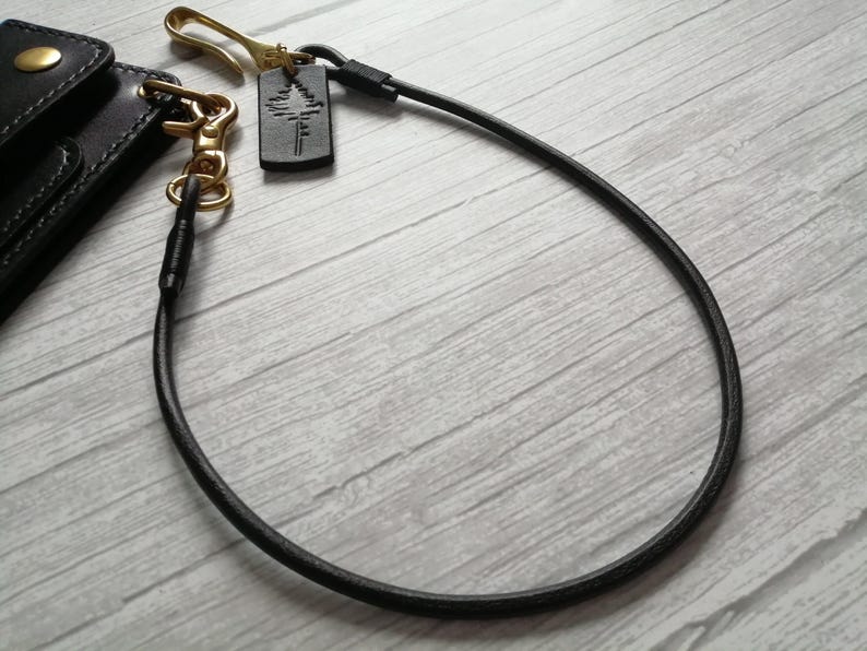 May include: A black leather wallet chain featuring a brass hook and a small rectangular leather tag embossed with a pine tree graphic. The chain connects to a black leather wallet corner with a brass snap button and a brass swivel lobster clasp.