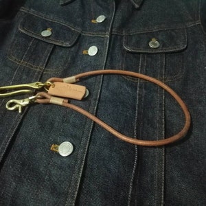 Puede incluir: Un cord&oacute;n de cuero marr&oacute;n para llaves con herrajes de lat&oacute;n, que incluye un cierre y un gancho. El cord&oacute;n se muestra sobre una chaqueta vaquera azul oscuro.