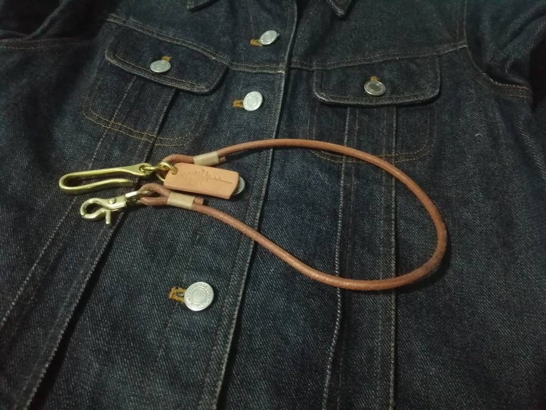 Puede incluir: Un cord&oacute;n de cuero marr&oacute;n para llaves con herrajes de lat&oacute;n, que incluye un cierre y un gancho. El cord&oacute;n se muestra sobre una chaqueta vaquera azul oscuro.
