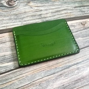 Puede incluir: Un porta tarjetas de cuero verde con un borde cosido y el texto "The Works Leathercraft" grabado en la parte delantera.