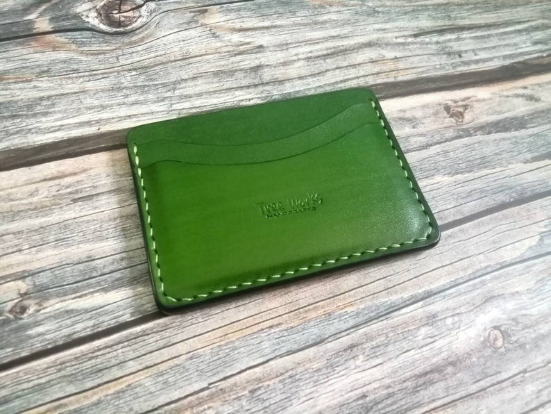 Puede incluir: Un porta tarjetas de cuero verde con un borde cosido y el texto "The Works Leathercraft" grabado en la parte delantera.