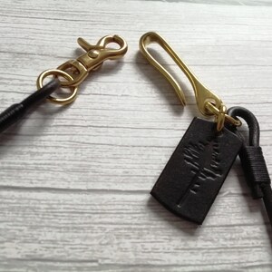 May include: A dark brown leather cord featuring a brass lobster claw clasp on one end and a brass hook clasp on the other. A small, rectangular dark brown leather tag with an embossed tree design is attached to the brass hook component.