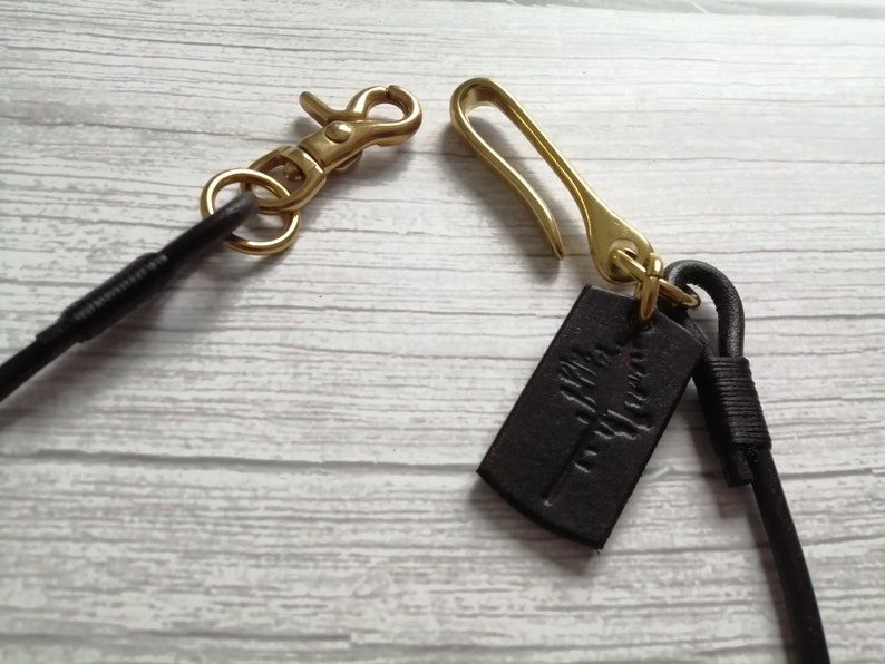 May include: A dark brown leather cord featuring a brass lobster claw clasp on one end and a brass hook clasp on the other. A small, rectangular dark brown leather tag with an embossed tree design is attached to the brass hook component.