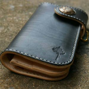 K&ouml;nnte beinhalten: Eine schwarze Ledergeldb&ouml;rse mit einem gesteppten Design und einem kleinen Baumlogo. Die Geldb&ouml;rse hat einen Klappverschluss mit einem Metallknopf.