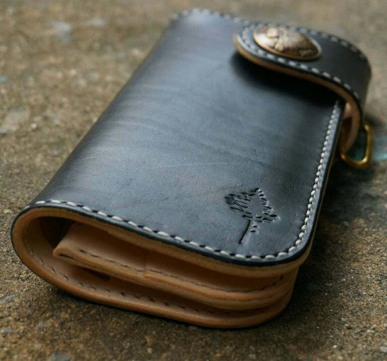 K&ouml;nnte beinhalten: Eine schwarze Ledergeldb&ouml;rse mit einem gesteppten Design und einem kleinen Baumlogo. Die Geldb&ouml;rse hat einen Klappverschluss mit einem Metallknopf.