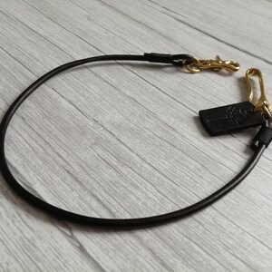 May include: A black leather cord strap featuring gold-toned metal hardware, including a swivel lobster clasp and a hook. A small black rectangular leather tag with an embossed tree logo is attached to the metal ring near the hook hardware.