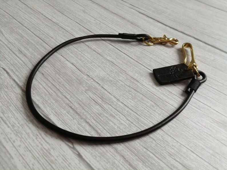 May include: A black leather cord strap featuring gold-toned metal hardware, including a swivel lobster clasp and a hook. A small black rectangular leather tag with an embossed tree logo is attached to the metal ring near the hook hardware.